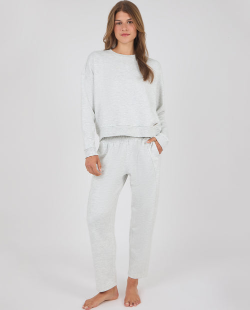 A woman modeling a light gray heathered lounge set consisting of a long-sleeve crewneck sweatshirt and matching jogger pants, standing against a plain light gray studio background.