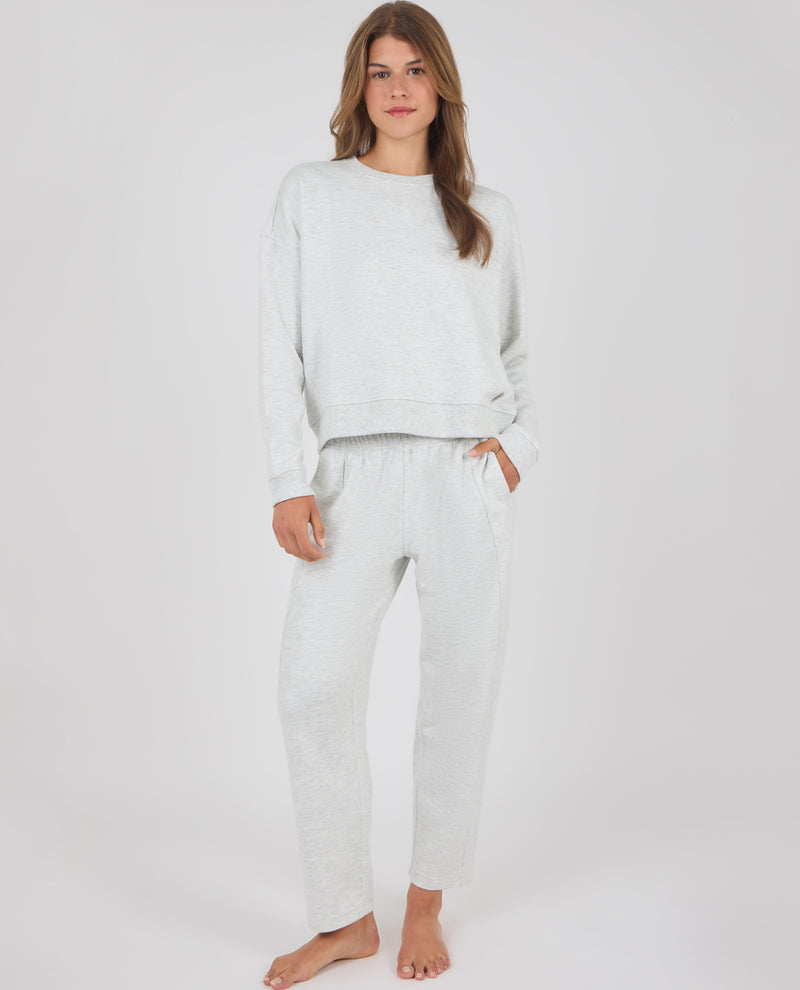 A woman modeling a light gray heathered lounge set consisting of a long-sleeve crewneck sweatshirt and matching jogger pants, standing against a plain light gray studio background.