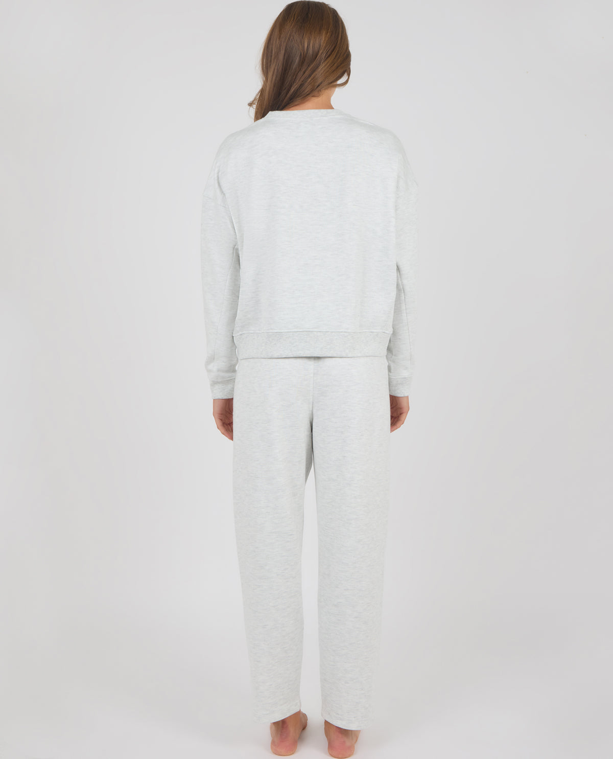A woman viewed from behind wearing a light heather gray matching lounge set consisting of a long-sleeve sweatshirt and wide-leg sweatpants, standing against a plain light gray studio background.