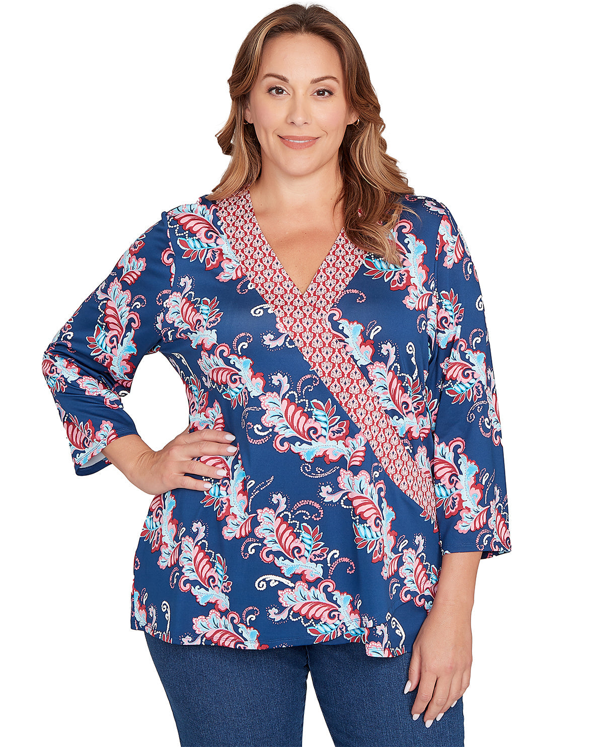A woman modeling a blue wrap-style blouse with a red and white paisley pattern and a red geometric patterned V-neck trim, paired with blue jeans against a plain white background.
