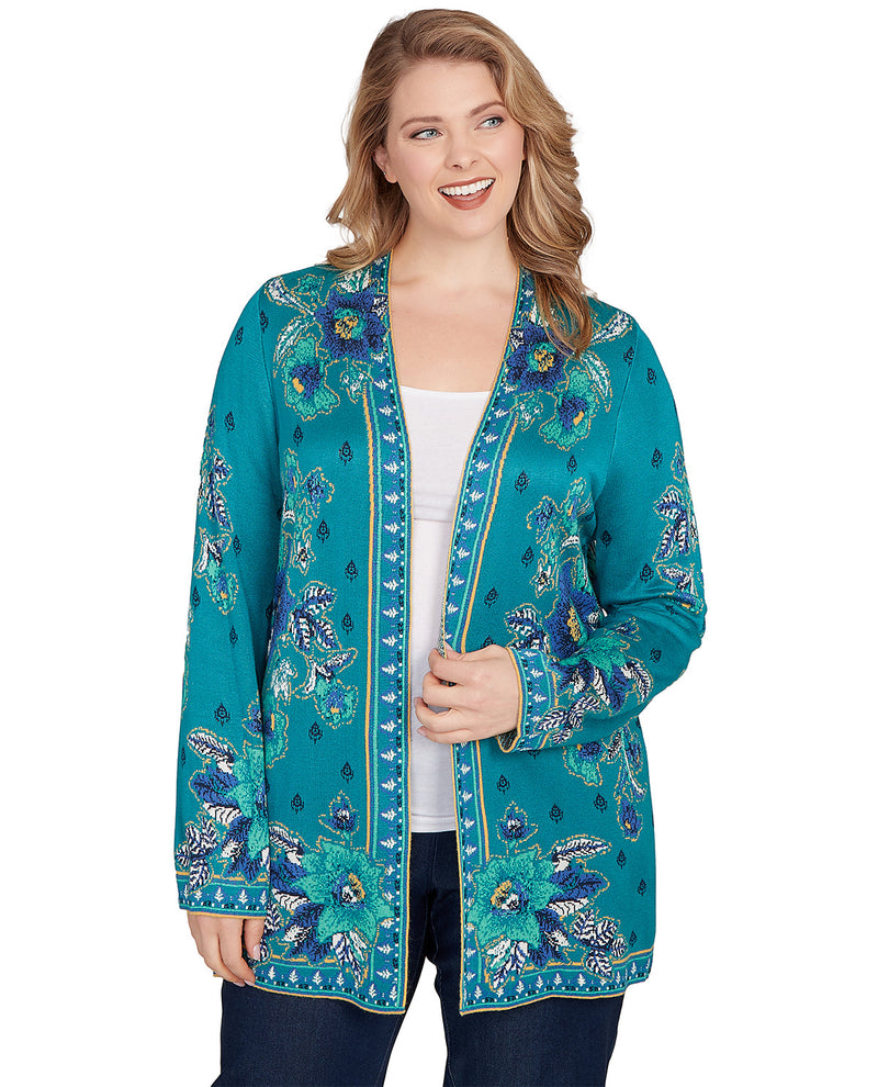A woman modeling a teal open-front cardigan with blue and white floral embroidery and gold trim, worn over a white top and dark jeans against a plain white background.