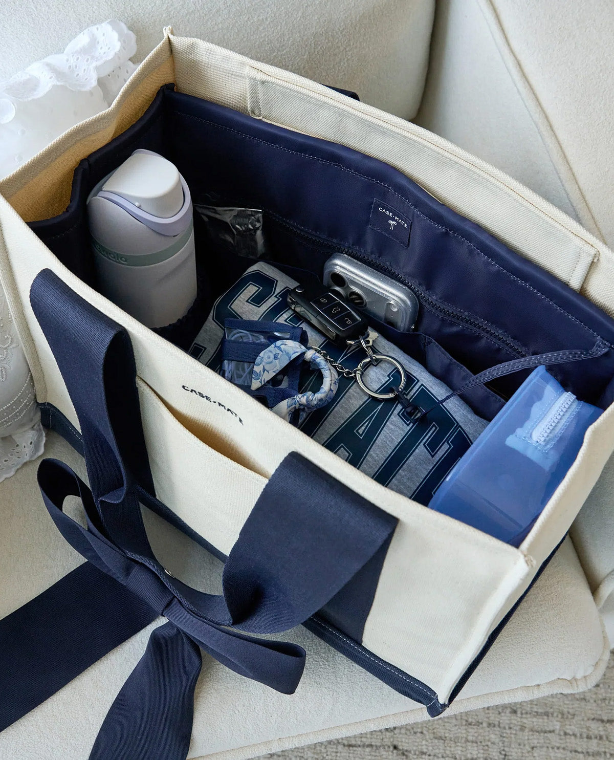 An open beige canvas organizer tote bag with navy ribbon handles, shown on a light upholstered chair. Inside are storage pockets holding a white bottle, a silver phone, keys, a patterned pouch, and a blue item.