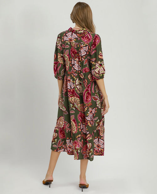 A woman viewed from the back wearing a knee-length floral dress with three-quarter sleeves. The dress has an olive green base with a large rose and floral print in pink, red, and beige tones.