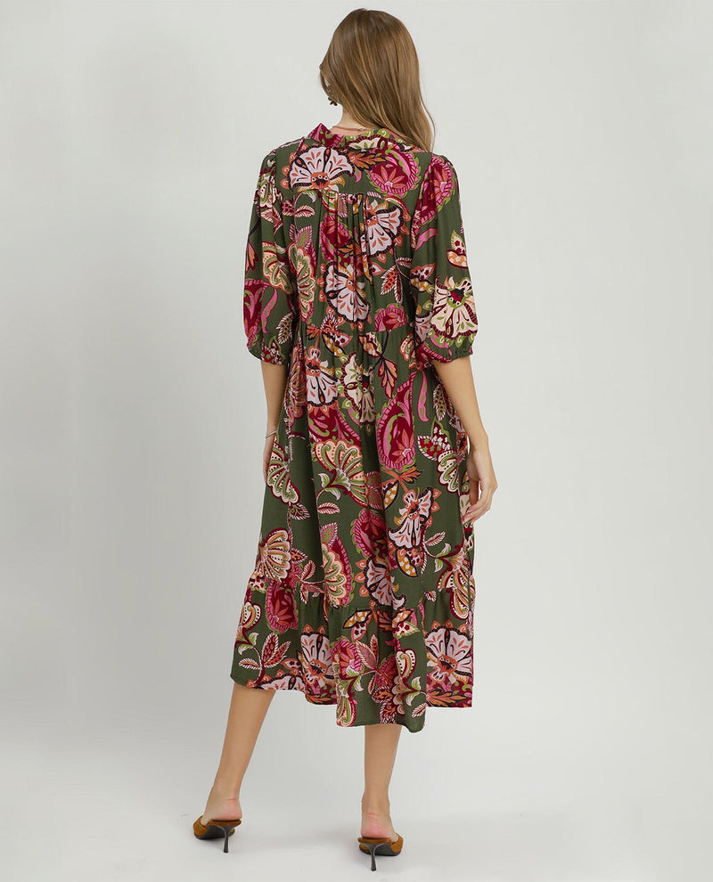 A woman viewed from the back wearing a knee-length floral dress with three-quarter sleeves. The dress has an olive green base with a large rose and floral print in pink, red, and beige tones.