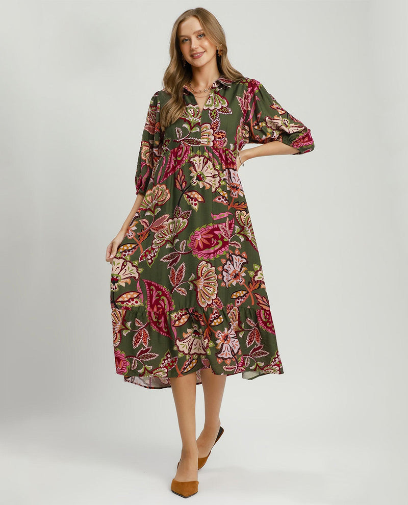 A woman modeling a knee-length midi dress with a dark olive green base and a vibrant floral print featuring pink, red, and beige roses and leaves, paired with brown pointed-toe heels against a light gray studio background.