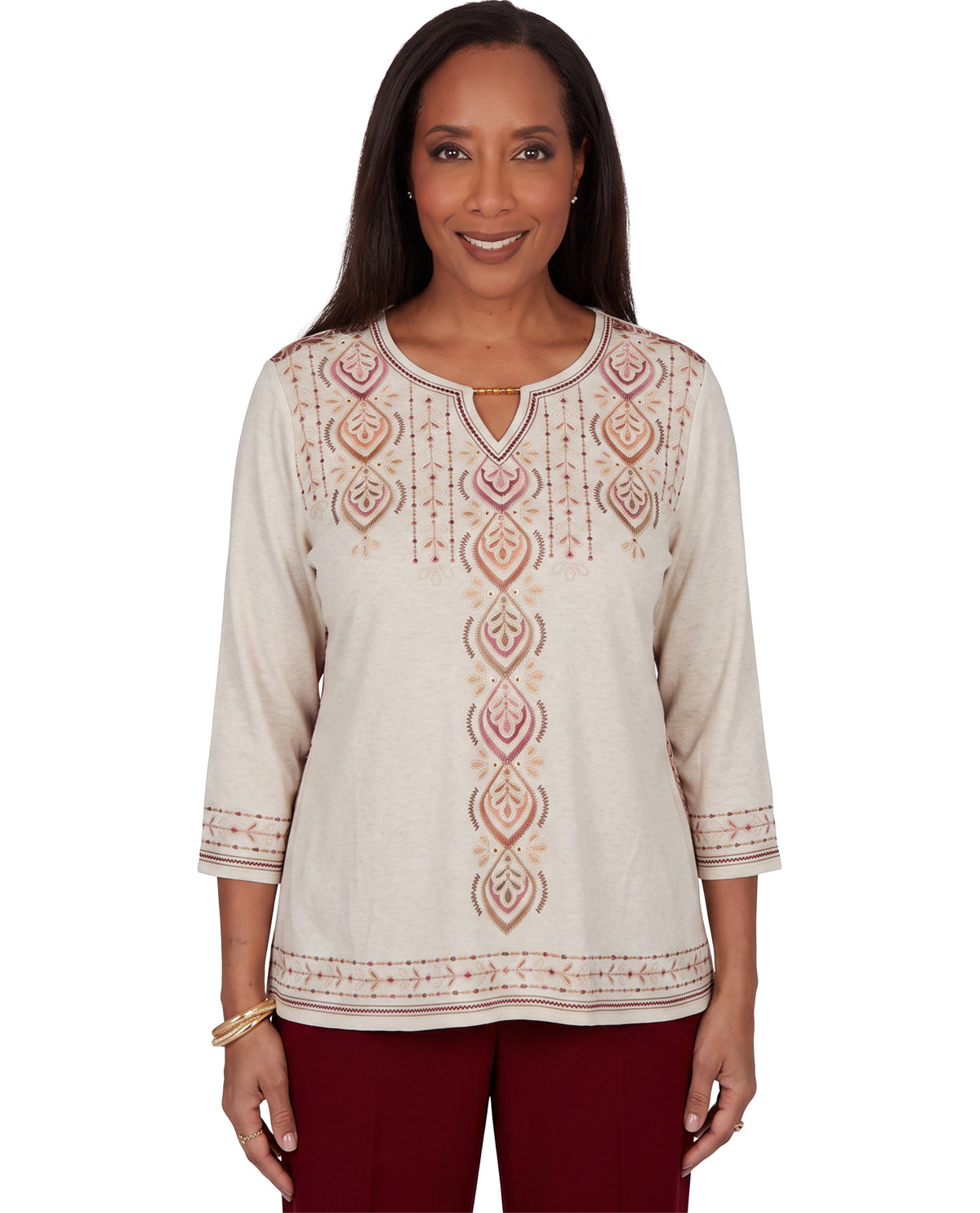 A woman modeling a cream-colored long-sleeve tunic top with pink and beige geometric embroidery, paired with burgundy pants, against a plain white background.