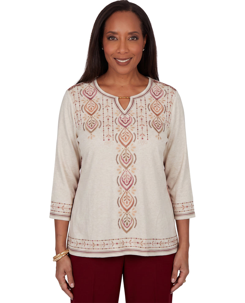 A woman modeling a cream-colored long-sleeve tunic top with pink and beige geometric embroidery, paired with burgundy pants, against a plain white background.