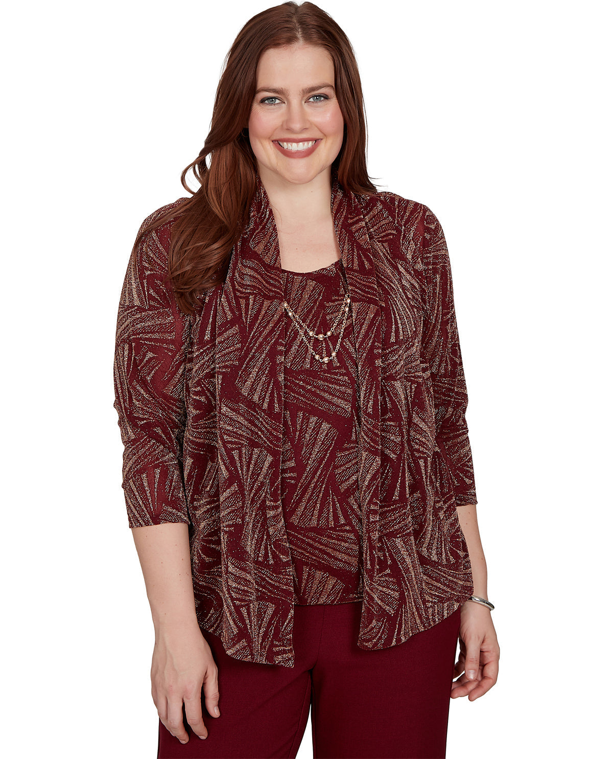 A woman modeling a rust red and beige abstract patterned blouse with three-quarter sleeves, paired with burgundy pants and layered necklaces, against a plain white background.