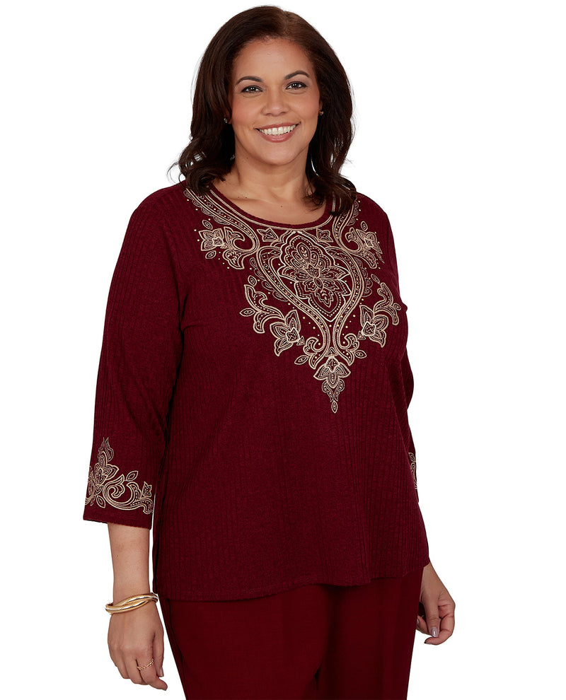 A woman wearing a burgundy three-quarter sleeve top with ornate beige embroidery on the chest and sleeve cuffs, paired with matching burgundy pants, against a plain white background.