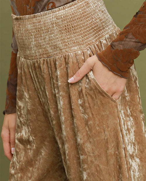 Close-up of a person wearing high-waisted, wide-leg trousers in a champagne gold crushed velvet fabric with a smocked waistband and side pockets.