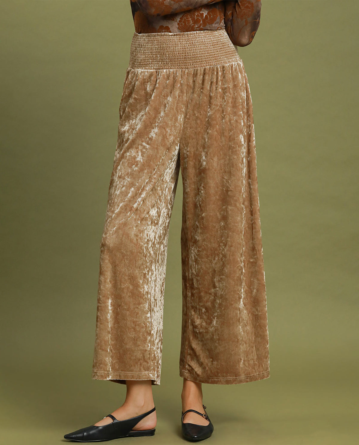 A person wearing high-waisted, wide-leg cropped pants in a brown champagne-colored velvet fabric with a textured elastic waistband, paired with black flat shoes, against a solid olive green studio background.