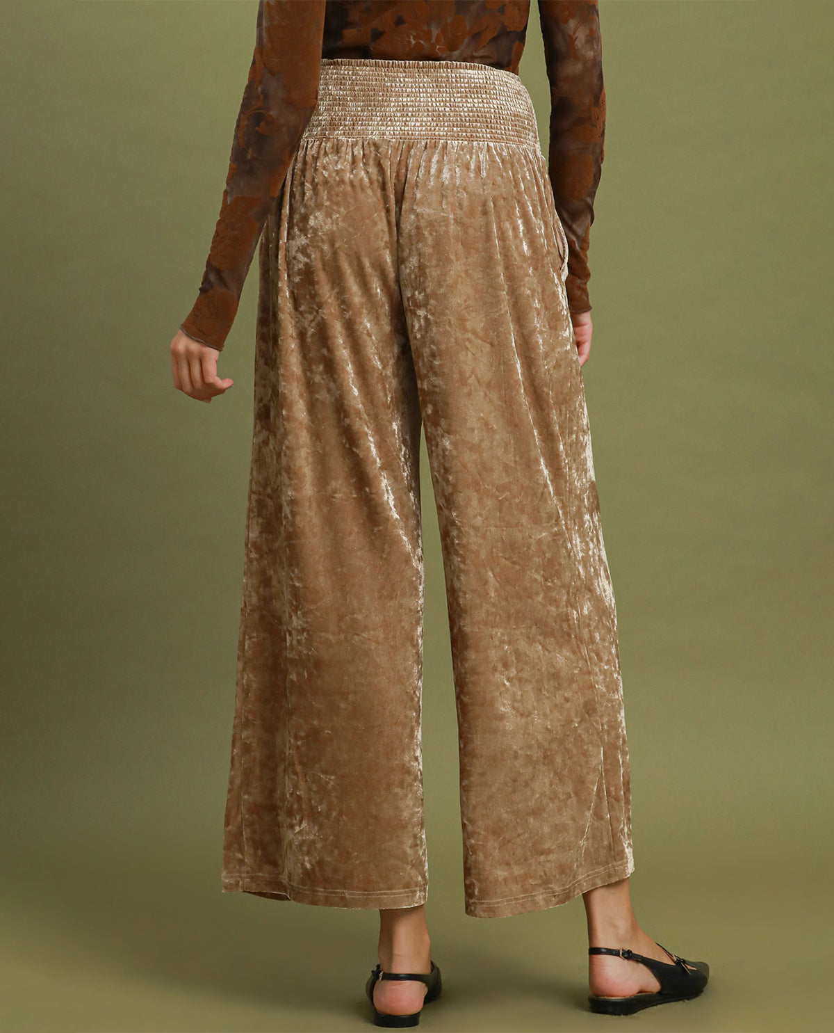 A model is shown from the back wearing high-waisted, wide-leg trousers in a textured, crushed brown velvet fabric. The background is a solid olive green studio backdrop.