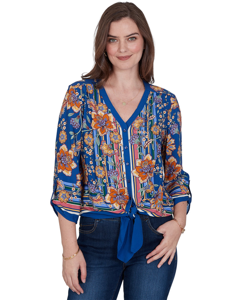 A woman modeling a blue floral and striped blouse with a V-neck and tie-front detail, paired with dark blue jeans, against a plain white background.
