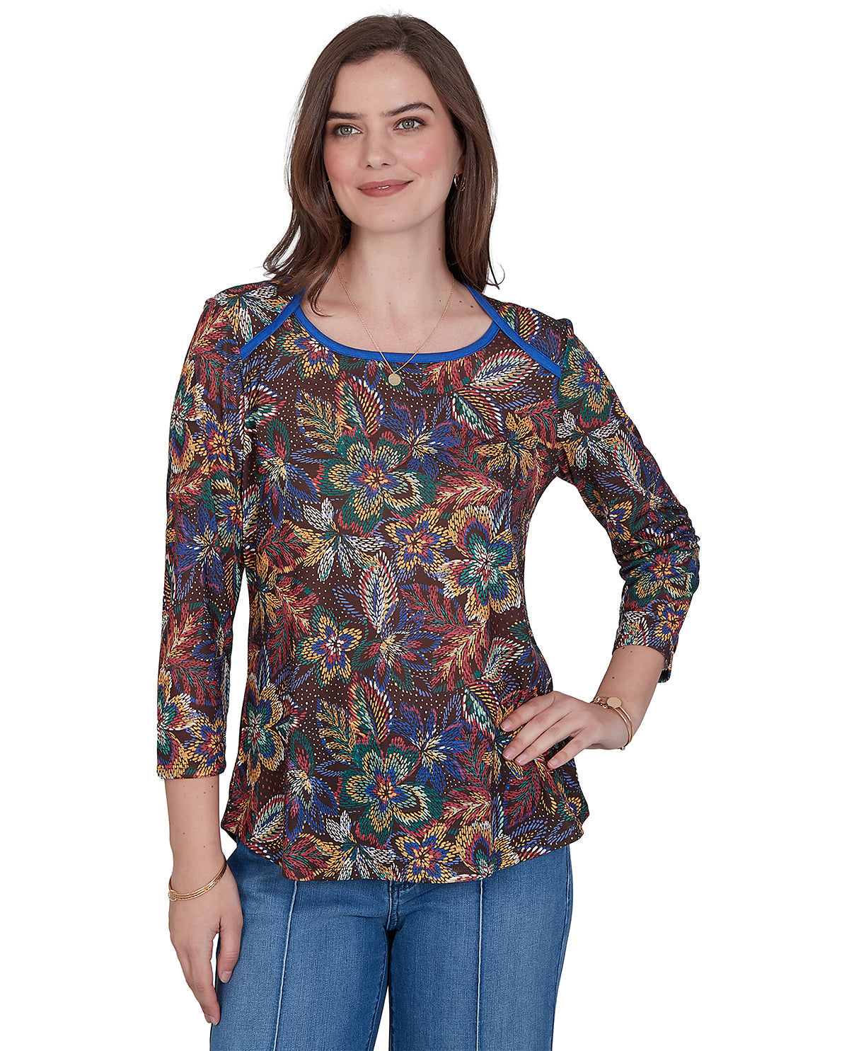 A woman modeling a three-quarter sleeve top with a dense multicolor floral and paisley print on a dark background, featuring a contrasting blue crew neck trim, paired with blue jeans against a plain white background.