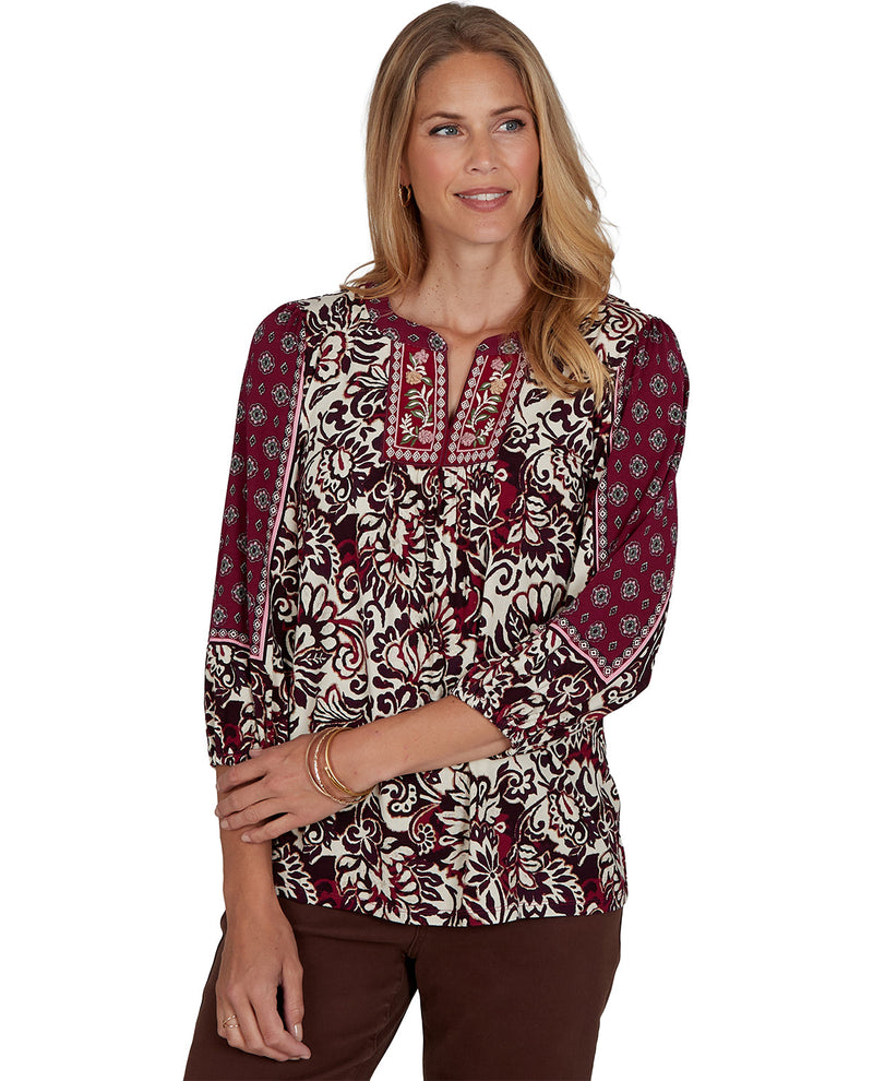 A woman modeling a long-sleeve blouse with a burgundy, cream, and brown floral and paisley pattern, paired with brown pants, against a plain white background.