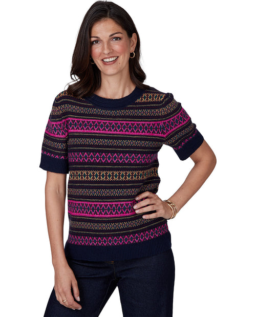 A woman modeling a short-sleeve crewneck knit sweater with horizontal geometric Fair Isle patterns in navy, magenta, gold, and black, paired with dark blue jeans against a plain white background.
