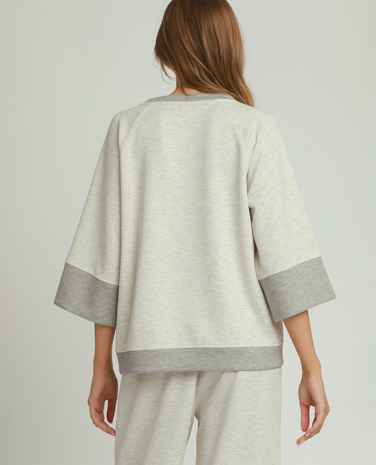 A woman shown from the back wearing a light heather gray sweatshirt with contrasting medium gray ribbed trim at the collar, sleeve cuffs, and hem, paired with matching light heather gray sweatpants, against a light gray studio background.