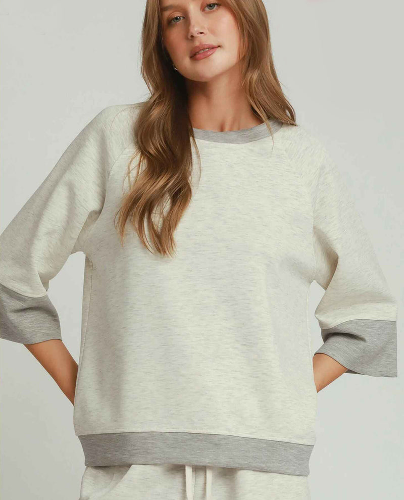A woman wearing a light heather gray sweatshirt with contrasting medium gray ribbed trim at the crew neck, sleeve cuffs, and hem, paired with matching light gray sweatpants against a solid light gray background.