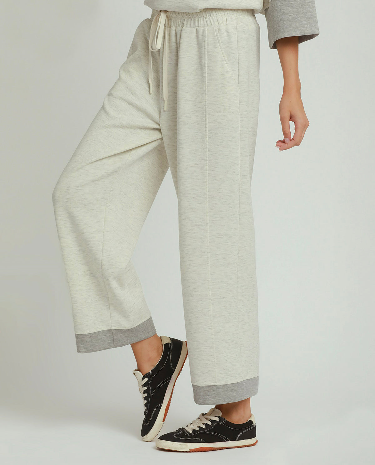 A person wearing light heather gray wide-leg cropped sweatpants with contrasting medium gray cuffs and a drawstring waist, paired with black low-top sneakers, against a light gray studio background.