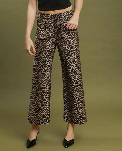 A model wearing high-waisted, wide-leg cropped pants with a brown and black leopard print, paired with a black crop top and black pointed-toe heels, set against a solid olive green studio background.