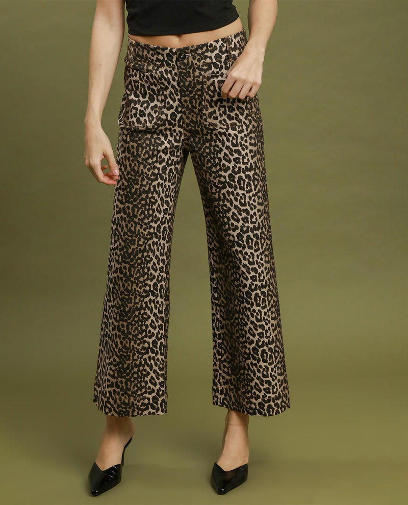 A model wearing high-waisted, wide-leg cropped pants with a brown and black leopard print, paired with a black crop top and black pointed-toe heels, set against a solid olive green studio background.