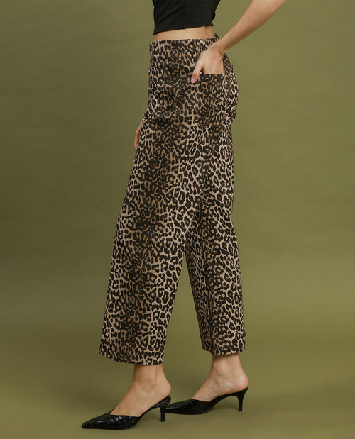 A model wearing high-waisted, wide-leg cropped pants with a brown and black leopard print, styled with black pointed-toe mule heels against a solid olive green studio background.