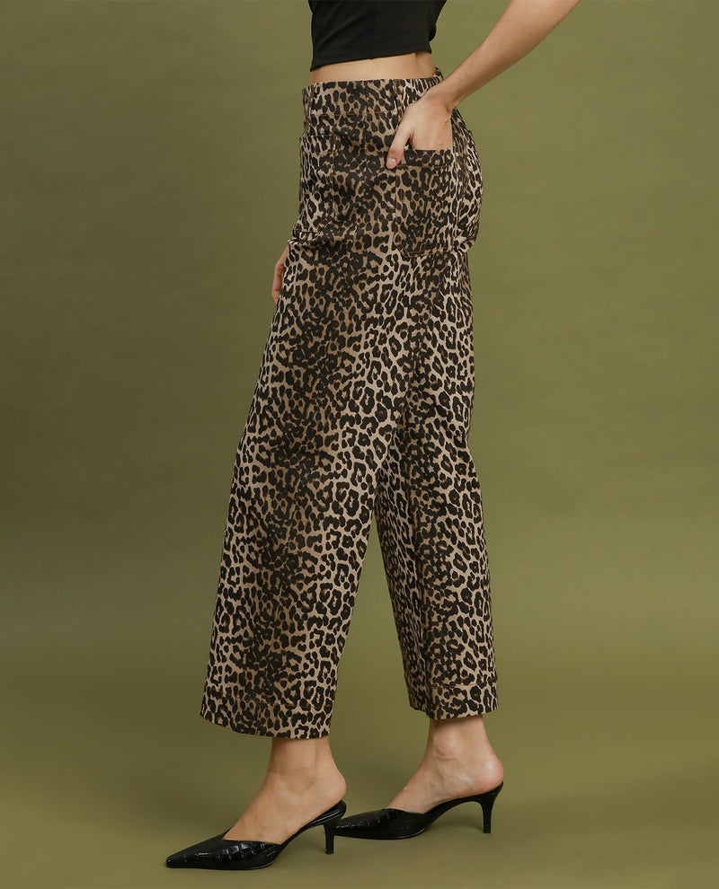 A model wearing high-waisted, wide-leg cropped pants with a brown and black leopard print, styled with black pointed-toe mule heels against a solid olive green studio background.