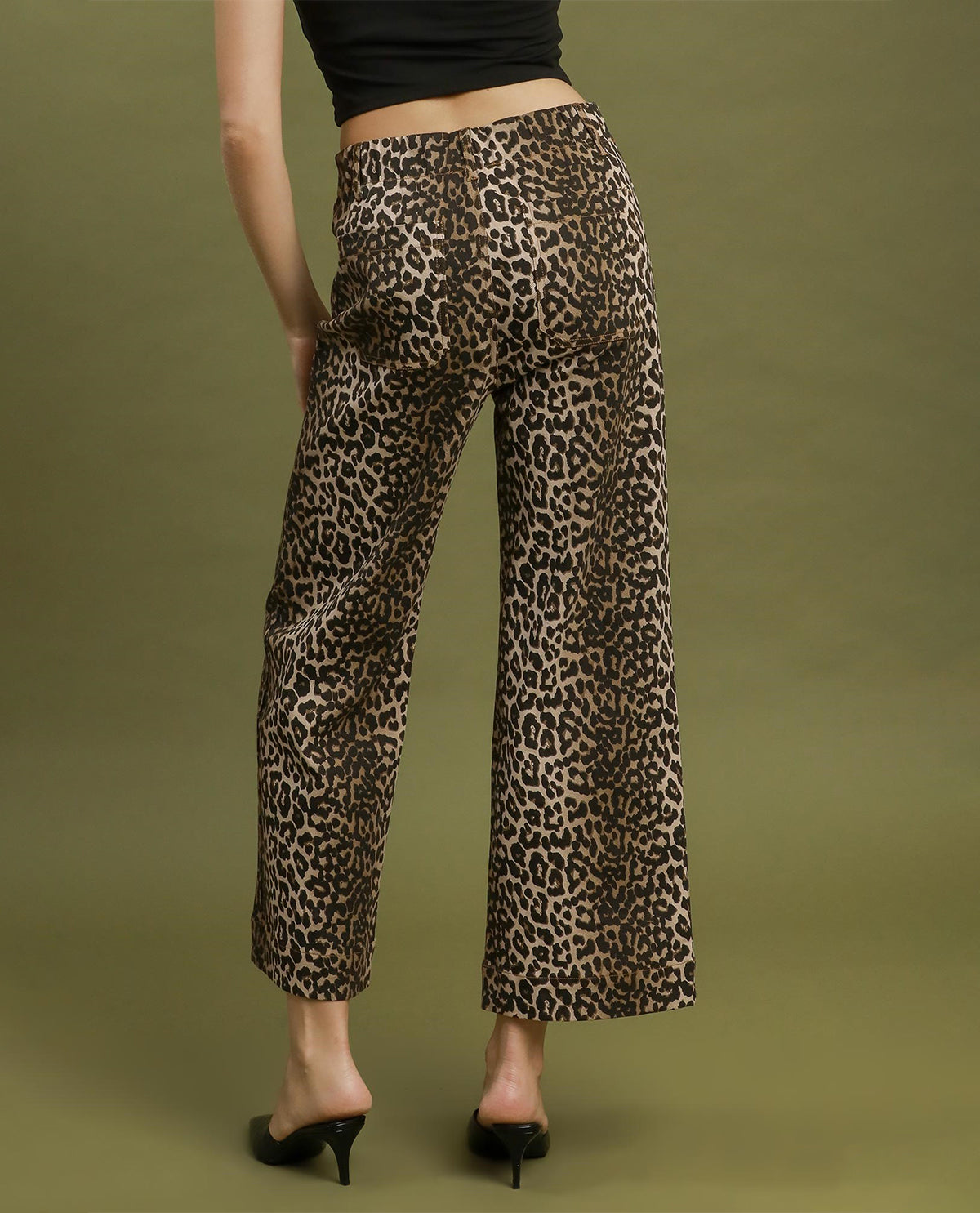 A model shown from the back wearing leopard-print wide-leg cropped pants, a black crop top, and black mule heels against a solid olive green studio background.