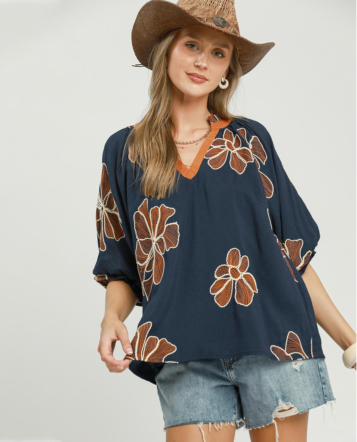 Woman wearing a navy blue floral-patterned top and denim shorts with a straw hat on a plain background