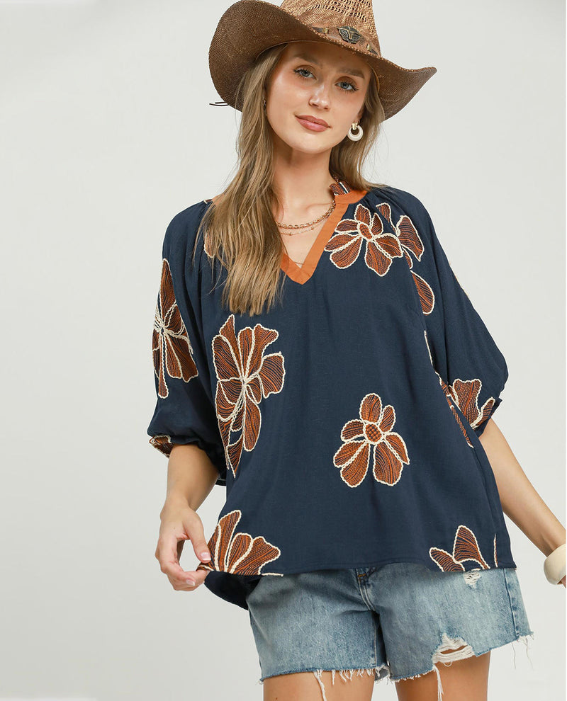 Woman wearing a navy blue floral-patterned top and denim shorts with a straw hat on a plain background