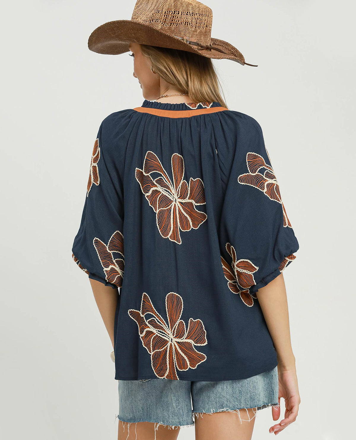 Woman wearing a navy blue blouse with brown floral patterns and a straw hat on a light gray background