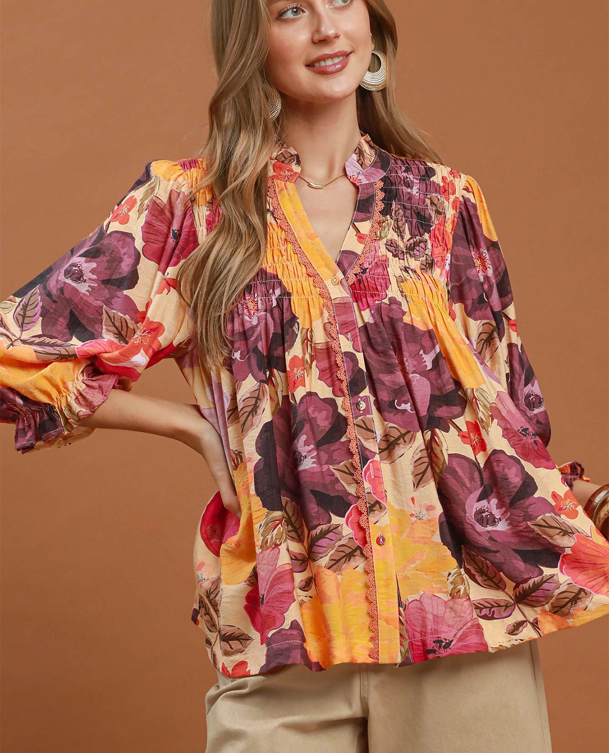 A woman wearing a vibrant floral print blouse with a mix of orange, purple, pink, and yellow tones, paired with beige pants, set against a solid warm brown background.