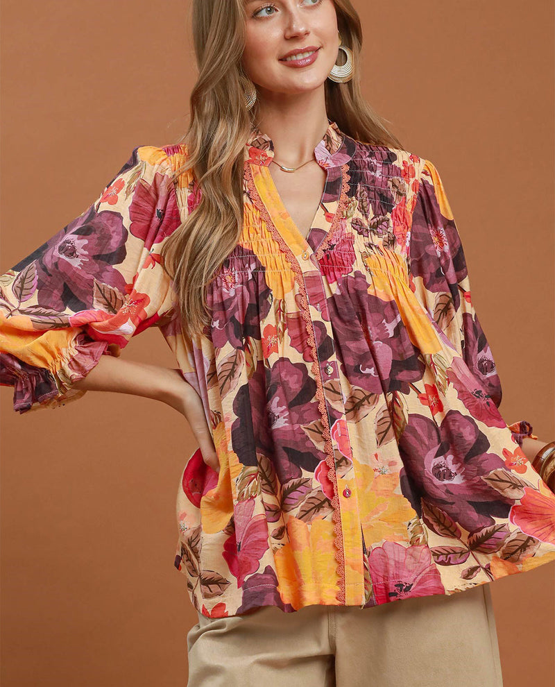 A woman wearing a vibrant floral print blouse with a mix of orange, purple, pink, and yellow tones, paired with beige pants, set against a solid warm brown background.