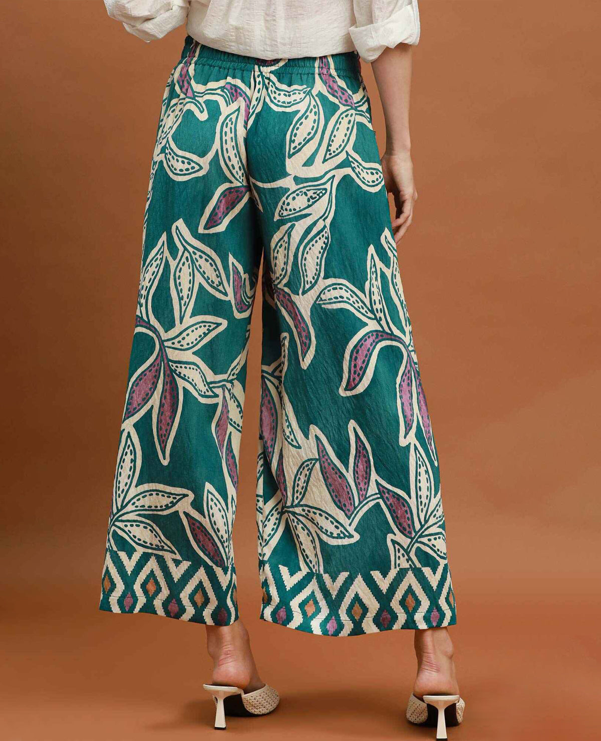 A person shown from the back wearing wide-leg cropped pants with a bold botanical print in teal, cream, and purple, paired with a white blouse and beige heeled sandals against a warm brown studio background.