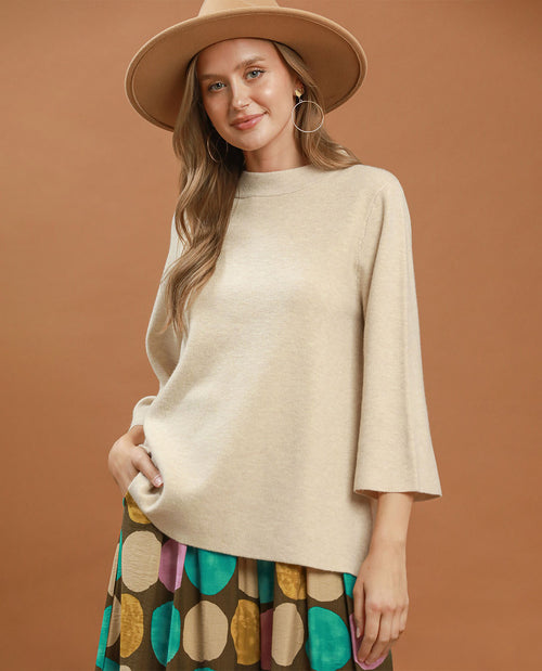 A woman wearing a cream-colored knit sweater with three-quarter bell sleeves, a wide-brimmed tan hat, and a colorful patterned skirt, posed against a solid warm brown studio background.