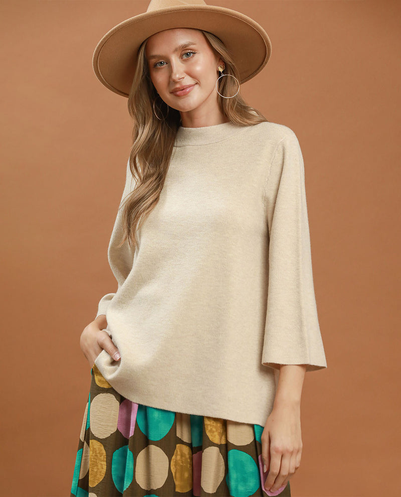 A woman wearing a cream-colored knit sweater with three-quarter bell sleeves, a wide-brimmed tan hat, and a colorful patterned skirt, posed against a solid warm brown studio background.