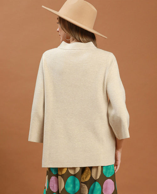 A woman shown from the back wearing a light beige knit blazer, a beige wide-brim hat, and a brown skirt with colorful polka dots, set against a solid terracotta background.