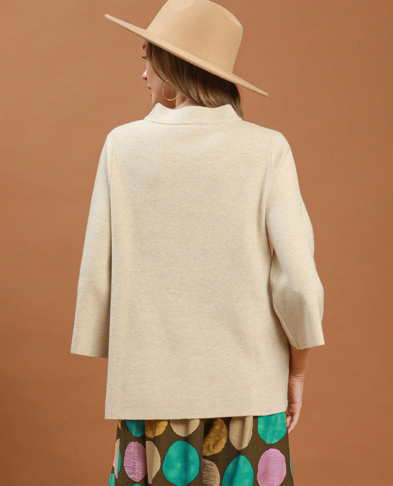 A woman shown from the back wearing a light beige knit blazer, a beige wide-brim hat, and a brown skirt with colorful polka dots, set against a solid terracotta background.