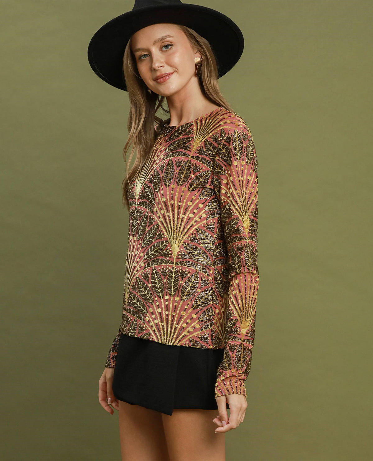 A woman wearing a long-sleeve knit top with a brown, pink, and gold geometric fan pattern, paired with black shorts and a black wide-brim hat, set against a solid olive green background.