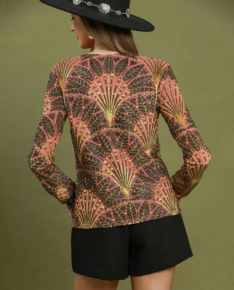A woman shown from the back wearing a long-sleeve top with a pink, gold, and dark brown art deco fan pattern, paired with black shorts and a black hat with silver embellishments, set against an olive green background.