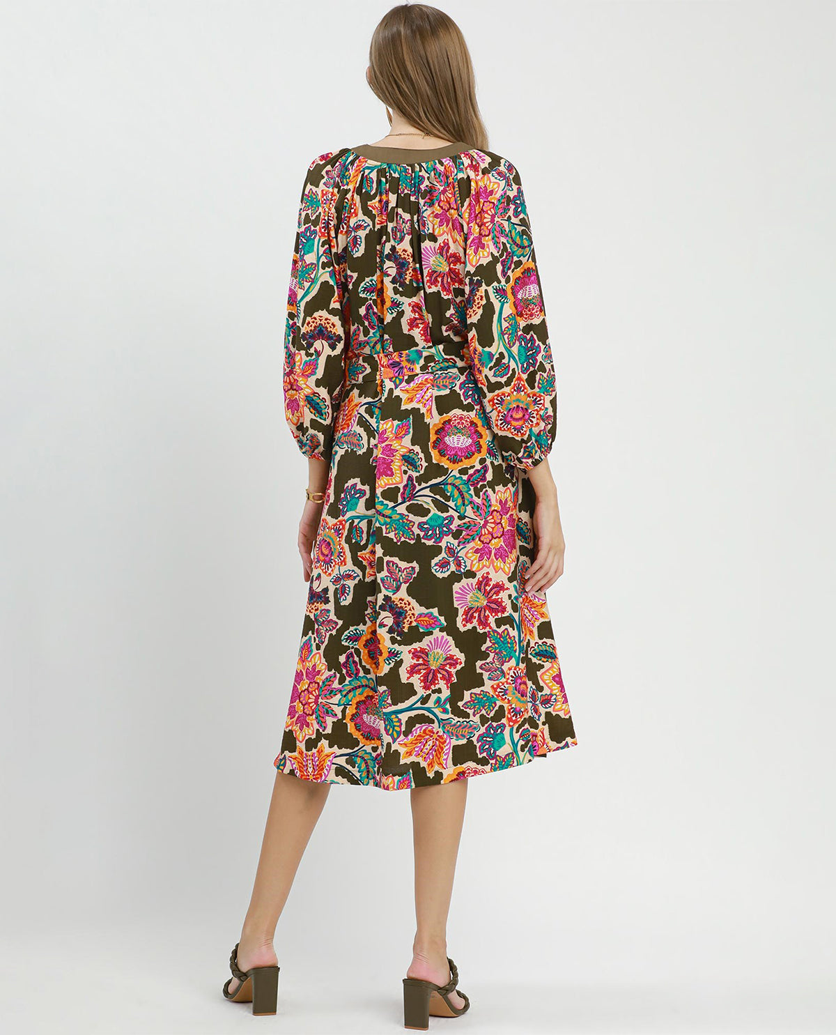 A woman viewed from the back wearing a long-sleeve midi dress with a dense multicolor floral print on a dark brown background, paired with brown block-heel sandals, against a light gray studio backdrop.