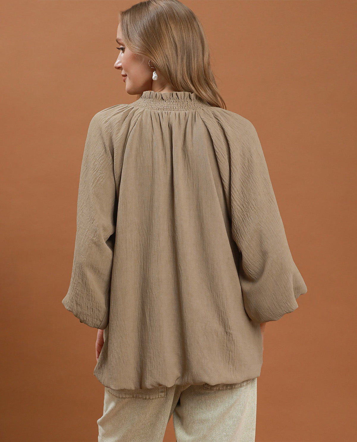 A woman shown from the back wearing a loose, textured taupe-beige blouse with a smocked neckline and voluminous sleeves, paired with light beige pants, against a solid warm brown studio background.