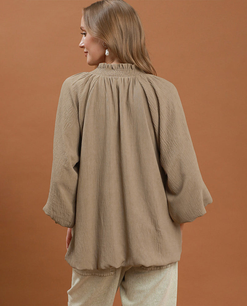 A woman shown from the back wearing a loose, textured taupe-beige blouse with a smocked neckline and voluminous sleeves, paired with light beige pants, against a solid warm brown studio background.