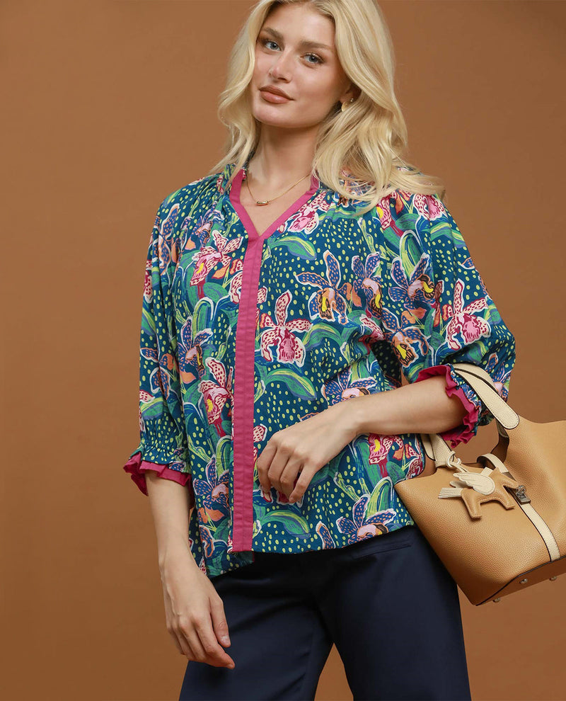 A woman models a vibrant, multicolored butterfly-print button-up blouse with pink ruffled trim, paired with navy pants and a tan handbag, set against a solid warm brown studio background.
