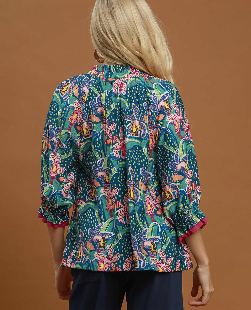A woman viewed from the back wearing a vibrant, multicolored tropical print blouse with three-quarter sleeves and dark navy pants, set against a solid warm brown studio background.