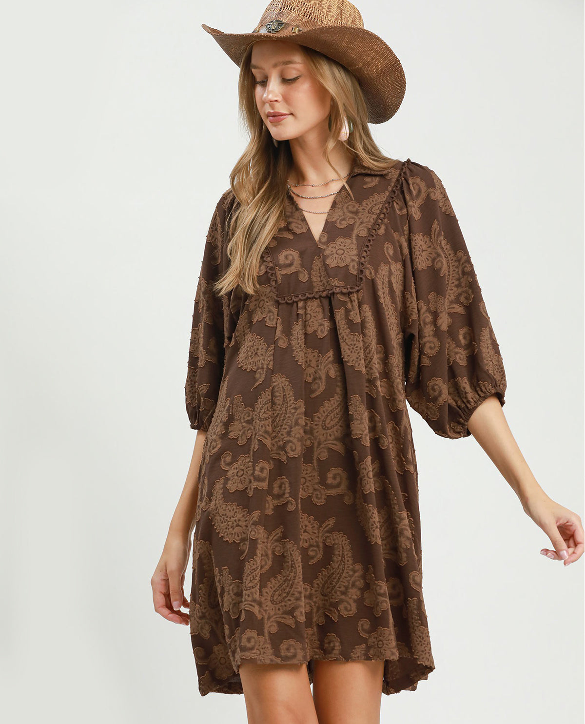 A woman wearing a brown bohemian-style mini dress with a paisley pattern, a cowboy hat, and layered necklaces, photographed against a light gray studio background.