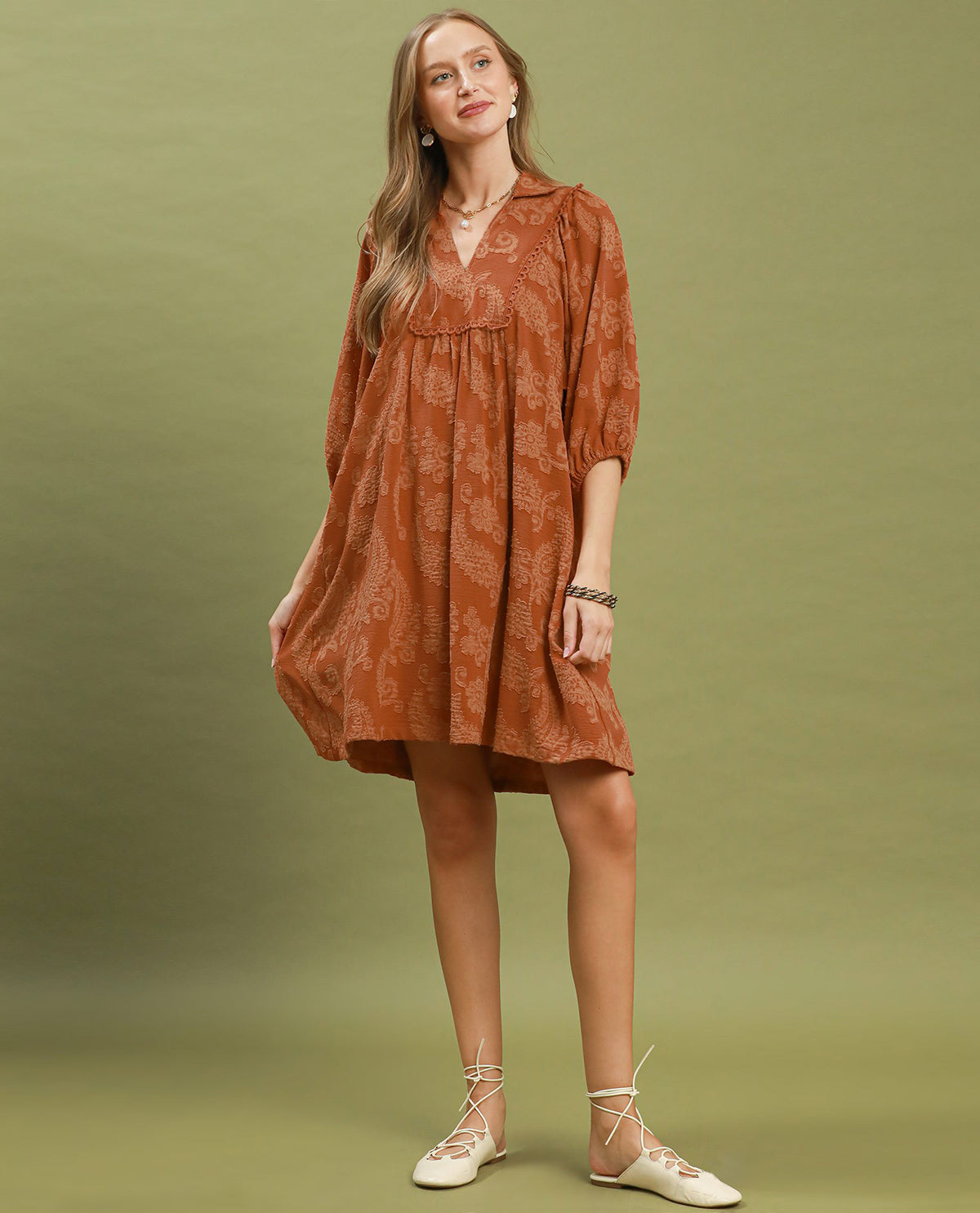 A woman modeling a rust-orange short-sleeve dress with a subtle paisley pattern, standing against a solid olive green studio background.