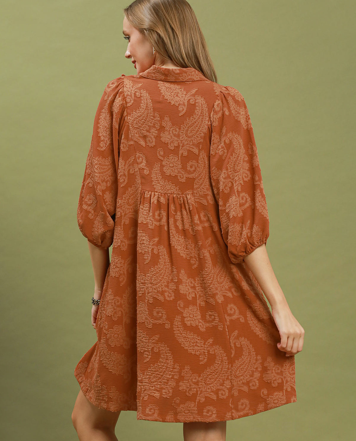 A woman wearing a rust-orange patterned tunic dress with three-quarter puff sleeves, shown from the back against a solid olive green background.