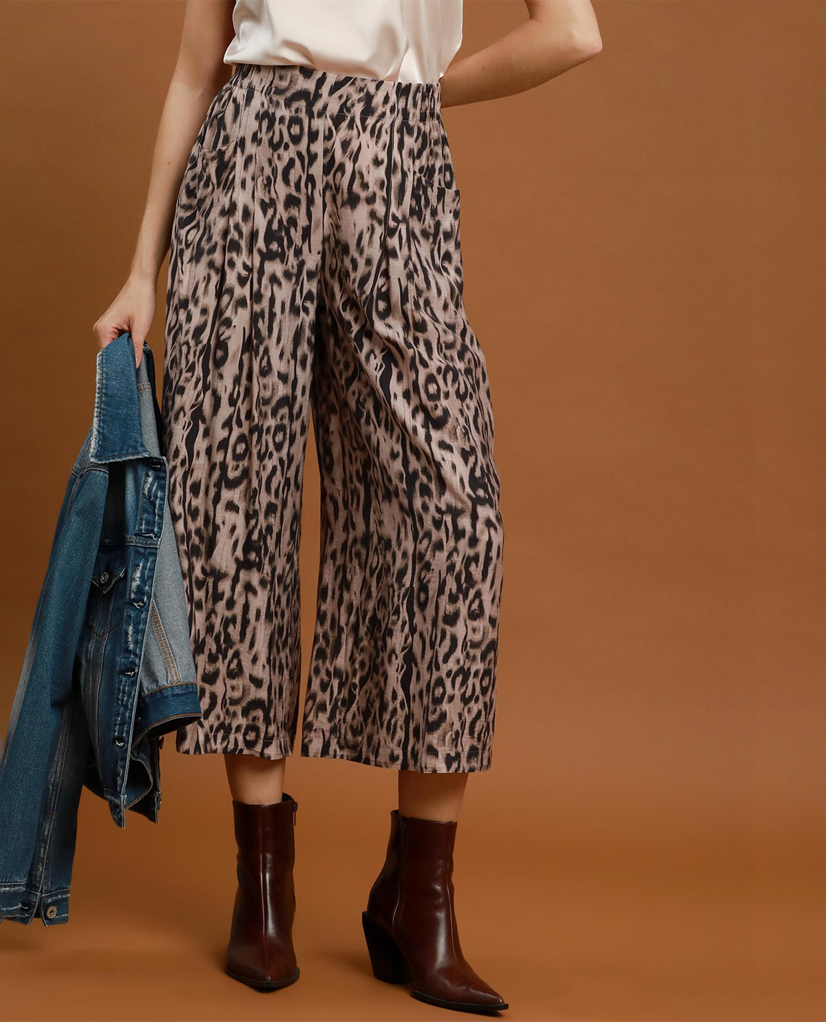 A model wearing high-waisted, wide-leg cropped pants with a beige, brown, and black leopard print pattern, styled with a white top, dark brown pointed-toe ankle boots, and holding a blue denim jacket against a solid warm brown studio background.