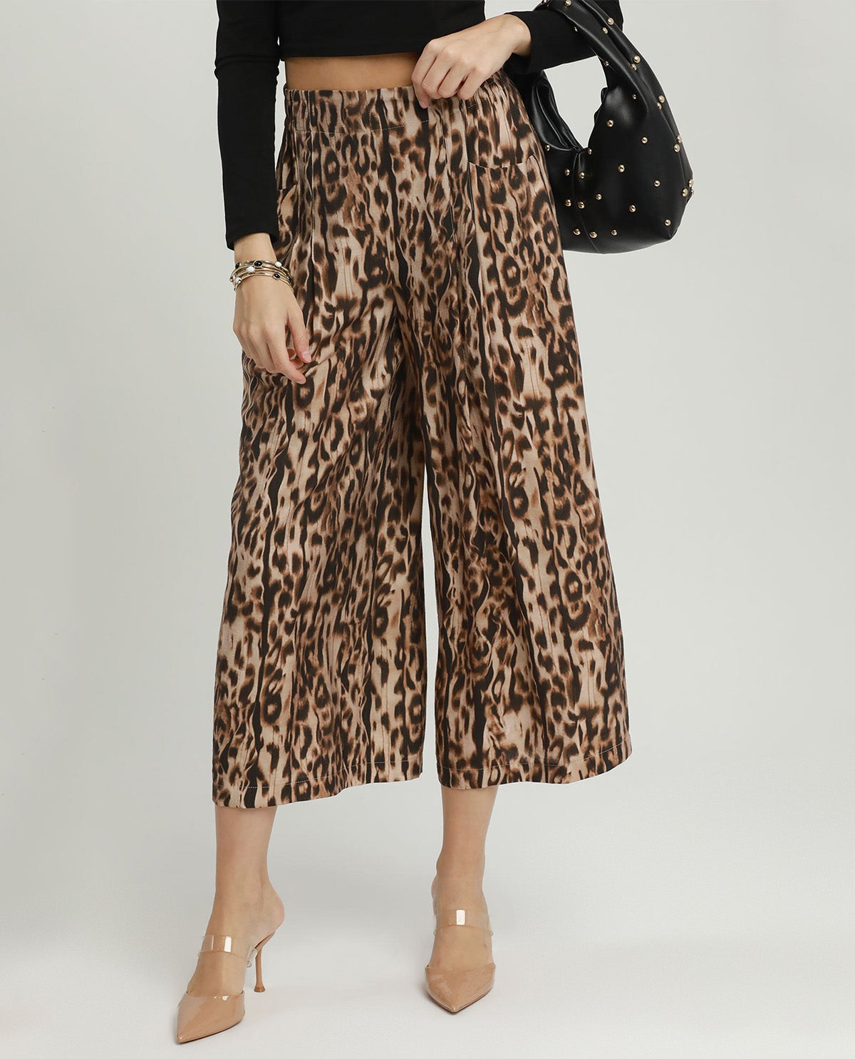 A model wearing high-waisted, wide-leg cropped pants with a brown and beige leopard print pattern, styled with a black long-sleeve crop top, beige high heels, and a black studded shoulder bag against a light gray studio background.